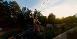 Человек едет на велосипеде по верёвочному мосту в парке «Паутина»